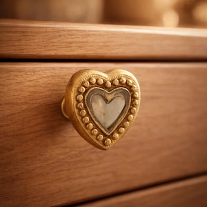 May include: A gold-colored heart-shaped drawer pull with a decorative beaded border and a central heart-shaped inlay. The drawer pull is mounted on a wooden drawer, showcasing a warm, natural wood grain.