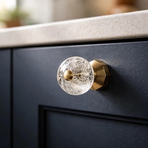 May include: A clear, crackled glass cabinet knob with a gold-colored base and center. The knob is round, and the base has a geometric design. The knob is mounted on a dark blue cabinet door.