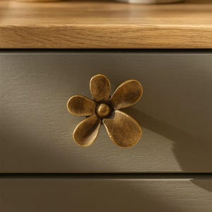 May include: A close-up of a drawer with a bronze-colored flower-shaped knob. The drawer is painted a muted green-gray color, and the top of the dresser is a light wood tone.