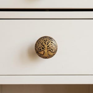 May include: A round, brass-colored drawer knob with a detailed tree of life design. The knob is mounted on a white drawer, offering a decorative and functional element for furniture. The tree design is intricately carved.
