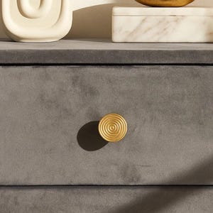 May include: Close-up of a gray velvet drawer with a gold spiral knob. The knob is centered on the drawer, casting a shadow. Other decorative items are visible in the background, including a white ceramic sculpture and a marble box.