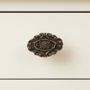 May include: An antique bronze-colored drawer pull with an ornate, oval design. The pull features a central floral motif surrounded by swirling, decorative elements. The background is a cream-colored drawer front.