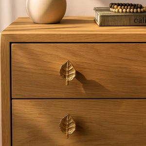 May include: Close-up of a wooden dresser with two drawers. Each drawer features a gold-colored leaf-shaped handle. The wood has a natural grain and a warm, light brown color. A decorative vase and a book are visible on top.