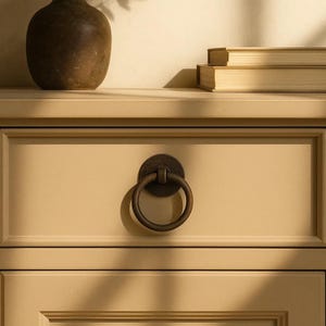 May include: A close-up of a beige drawer with a dark bronze ring pull. The drawer is part of a larger piece of furniture, with a vase and books visible on the top surface. The ring pull is circular and attached to a circular backplate.