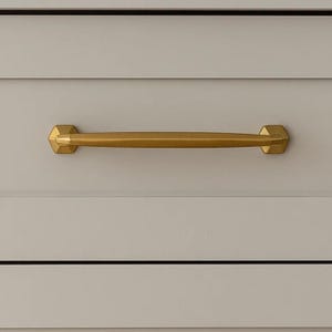 May include: A gold-colored cabinet pull with a geometric design. The pull is a horizontal bar with hexagonal ends, mounted on a light gray cabinet door. The hardware adds a touch of elegance to the cabinetry.