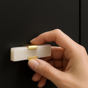 May include: A rectangular cabinet pull with a white marble body and gold-colored metal accents. The hardware is being held against a black cabinet door. The pull is designed for easy gripping and opening of drawers or cabinets.
