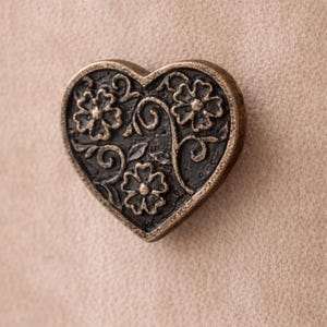 May include: A heart-shaped decorative knob with an antique bronze finish. The knob features a floral design with three flowers and swirling vines. The background is a neutral beige color.