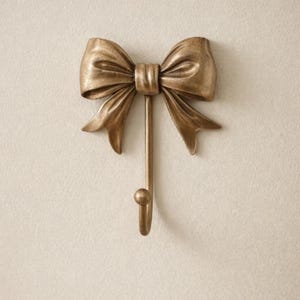May include: A gold-colored metal wall hook shaped like a bow. The bow has detailed folds and a central knot, with a long, straight hook extending downwards. The hook ends in a small, rounded ball.