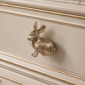 May include: A gold-colored bunny-shaped drawer pull mounted on a light gray drawer front. The drawer front has gold trim and a recessed panel design. The bunny pull is detailed with visible fur and a sitting pose.
