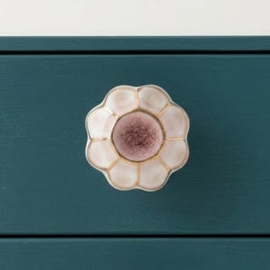 May include: A flower-shaped drawer pull with a white and pink color scheme and gold accents. The drawer pull is set against a teal-colored drawer front, creating a contrast. This decorative hardware adds a touch of elegance.