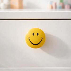 May include: A yellow smiley face cabinet knob with a black smile and two black eyes. The knob is round and mounted on a white cabinet door. The knob adds a cheerful touch to any furniture.