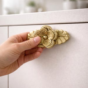 May include: A gold-colored cabinet pull with a floral design. The pull features a central flower shape with detailed petals and is flanked by leaf-shaped accents. The hardware is mounted on a white cabinet door.