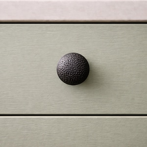 May include: Close-up of a dark, textured, round cabinet knob. The knob is centered on a light green drawer front with a horizontal wood grain pattern. A white surface is above the drawer.