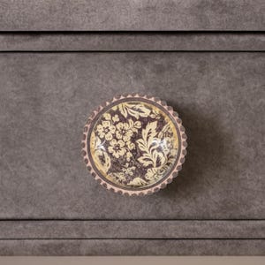 May include: A round cabinet knob with a floral design. The knob features a cream-colored floral pattern on a dark purple background, encased in a scalloped edge. The knob is set against a gray cabinet.