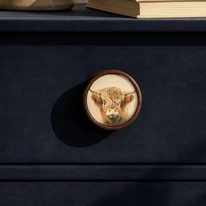 May include: A round wooden cabinet knob featuring a detailed image of a Highland cow. The knob has a brown wooden frame and a light beige background. The cow has long, shaggy, light brown fur and large horns. The knob is mounted on a dark blue cabinet.