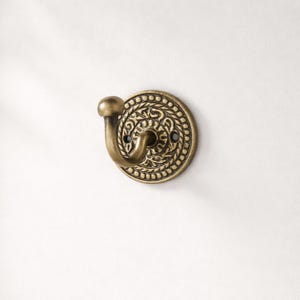 May include: A decorative brass wall hook with an ornate, circular design. The hook features a rounded knob and a curved hook, attached to a circular base with a detailed pattern. The hook is mounted on a white surface.