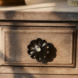 May include: Close-up of a drawer with a dark, flower-shaped knob. The knob has a central crystal accent. The drawer is a muted brown color, with a recessed panel and a soft, velvety texture. The overall aesthetic is elegant and refined.
