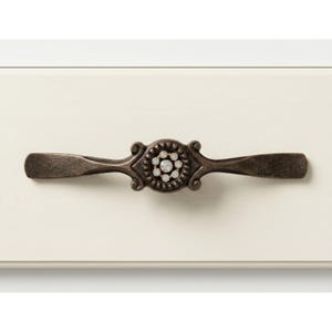 May include: A bronze-toned cabinet pull with a central floral design encrusted with small, clear crystals. The pull has a symmetrical design with elongated, rectangular handles on each end. The hardware is set against a cream-colored cabinet surface.