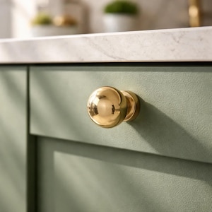 May include: A close-up of a gold-colored cabinet knob with a round shape. The knob is mounted on a light green cabinet door. The background shows a white countertop and a glimpse of plants.