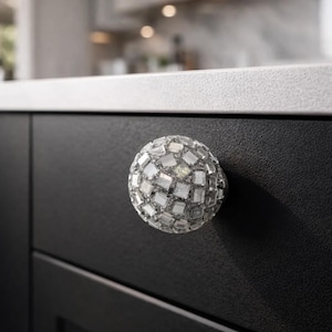 May include: A decorative cabinet knob with a spherical shape. The knob is covered in small, reflective, square-shaped pieces, creating a mosaic-like appearance. It is mounted on a black cabinet door, offering a contrasting visual element.