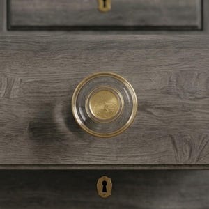 May include: Close-up of a gray wooden cabinet with a round, clear glass and gold-toned drawer pull. The pull has a gold center with a design and a gold rim. A gold-toned keyhole is visible below the drawer pull.