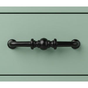 May include: Close-up of a black metal drawer pull with a decorative design. The pull is centered on a light green drawer front. The pull has a central sphere and decorative detailing on either side.