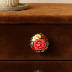 May include: A decorative, round cabinet knob with a floral design. The knob features a red flower at the center, surrounded by yellow, blue, and white petals. The knob is set against a dark brown cabinet.