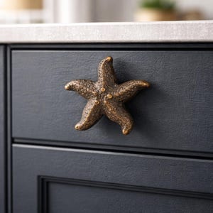May include: Bronze starfish cabinet knob. The five-pointed knob is mounted on a dark blue cabinet door. The knob has a textured surface and is a decorative accent for coastal or nautical themed decor.