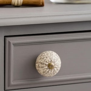 May include: A gray painted drawer with a round, speckled ceramic knob. The knob has a brass-colored base and a pattern of white and gray spots and lines. The drawer is part of a gray cabinet.