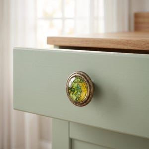 May include: A round drawer knob featuring a vintage map design under a clear glass dome. The knob has a distressed, antique bronze-colored metal frame and is mounted on a light sage green wooden drawer front with a natural wood top edge.