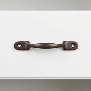 May include: A bronze-colored drawer pull with a curved handle and two mounting holes. The hardware is set against a white surface, providing a contrast that highlights the pull's design. This piece is suitable for cabinets and drawers.