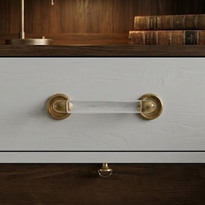 May include: A clear acrylic and brass drawer pull is centered on a white drawer. The brass hardware has a circular design. The drawer pull is a cylinder shape. The drawer is above a dark brown drawer with a matching brass knob.