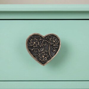 May include: A heart-shaped drawer pull with a floral design in antique bronze finish. The drawer pull is mounted on a light teal painted surface. The floral design includes flowers and swirling vines.
