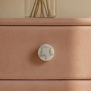 May include: A close-up of a drawer with a round, white and gray marble-patterned knob. The drawer is a muted rose color. A glass vase with dried stems is visible in the background.