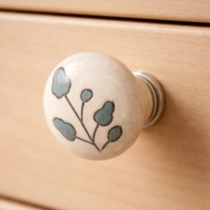 May include: A cream-colored ceramic cabinet knob with a floral design. The knob features a round shape with a silver metal base. The floral design includes teal-colored leaves and a black stem.