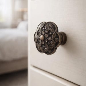 May include: Close-up of a decorative bronze-colored drawer pull. The pull is shaped like a flower with intricate leaf and petal details. The knob is mounted on a cream-colored cabinet door, suggesting a vintage or antique style.