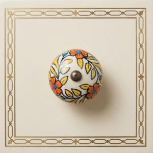May include: A round, white ceramic cabinet knob with a floral design in orange, blue, and yellow. The knob has a silver-colored metal screw and is set against a cream-colored square with a gold geometric border.