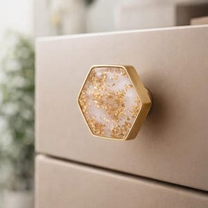 May include: A gold-toned, hexagonal cabinet knob with a white and gold flecked center. The knob is mounted on a light brown cabinet drawer. The knob's design adds a touch of elegance to furniture.