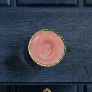 May include: A round cabinet knob with a pink and white swirl design, framed by a gold-colored edge. The knob is mounted on a dark blue wooden surface, showcasing a decorative hardware element.