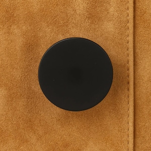 May include: A close-up of a black, circular button on a textured, tan fabric. The button is centered and appears to be made of a smooth, matte material. The fabric has a suede-like appearance.
