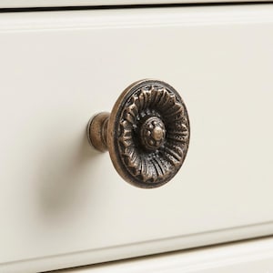 May include: Close-up of a bronze-colored drawer pull with a floral design. The round pull is mounted on a cream-colored drawer front. The pull has a detailed, textured surface and a small, central knob.