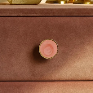 May include: A close-up of a pink and gold cabinet knob. The knob has a circular design with a pink center and a gold-colored rim. The knob is mounted on a dusty rose-colored cabinet drawer.