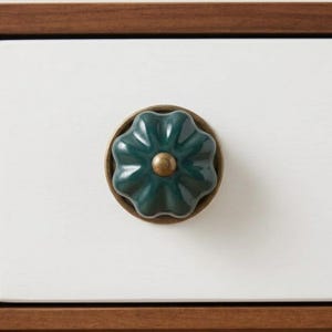 May include: A dark teal flower-shaped cabinet knob with a brass base and center. The knob is mounted on a white drawer with a dark wood trim. The design is a decorative accent for furniture.