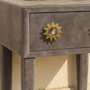 May include: A gray velvet nightstand with a drawer featuring a gold starburst knob with a clear glass center. The nightstand has a rectangular shape and four legs.