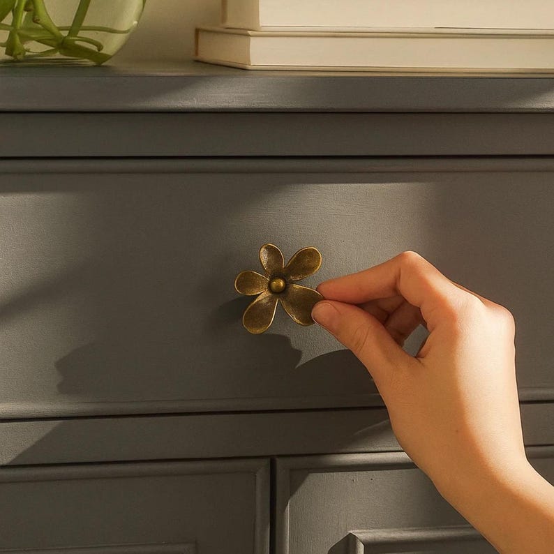 May include: A close-up of a flower-shaped cabinet knob in antique brass finish. The knob is being held by a hand against a gray cabinet door. The knob has five petals and a central circular detail.