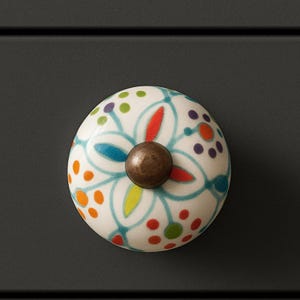 May include: A decorative ceramic cabinet knob with a floral design. The knob is white with a pattern of colorful dots and a central flower in blue, red, and yellow. It has a bronze-colored metal base.