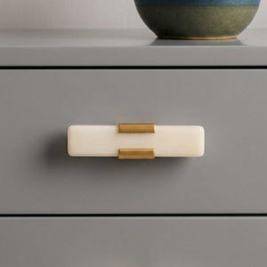 May include: A rectangular, off-white cabinet pull with brass-colored accents. The pull is mounted on a gray cabinet drawer. A ceramic bowl with blue and green tones sits on top of the cabinet.