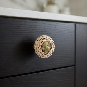 May include: A decorative cabinet knob with a woven, light beige exterior and a central, antique brass-colored medallion. The knob is mounted on a dark brown cabinet door, offering a rustic yet elegant aesthetic.