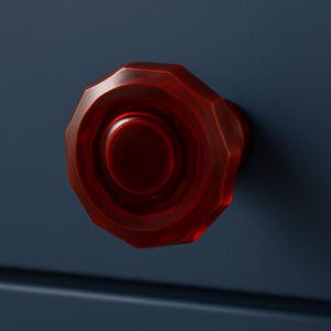 May include: A close-up of a red, faceted cabinet knob. The knob is a deep, translucent red with a circular center and an octagonal outer shape. The knob is mounted on a dark blue surface.