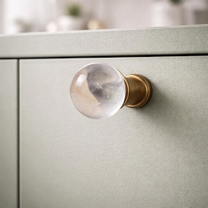 May include: A clear, spherical cabinet knob with a brass base is attached to a light green cabinet door. The knob has a marbled, cloudy appearance within the clear glass. The brass base is circular and provides a contrasting accent to the knob.
